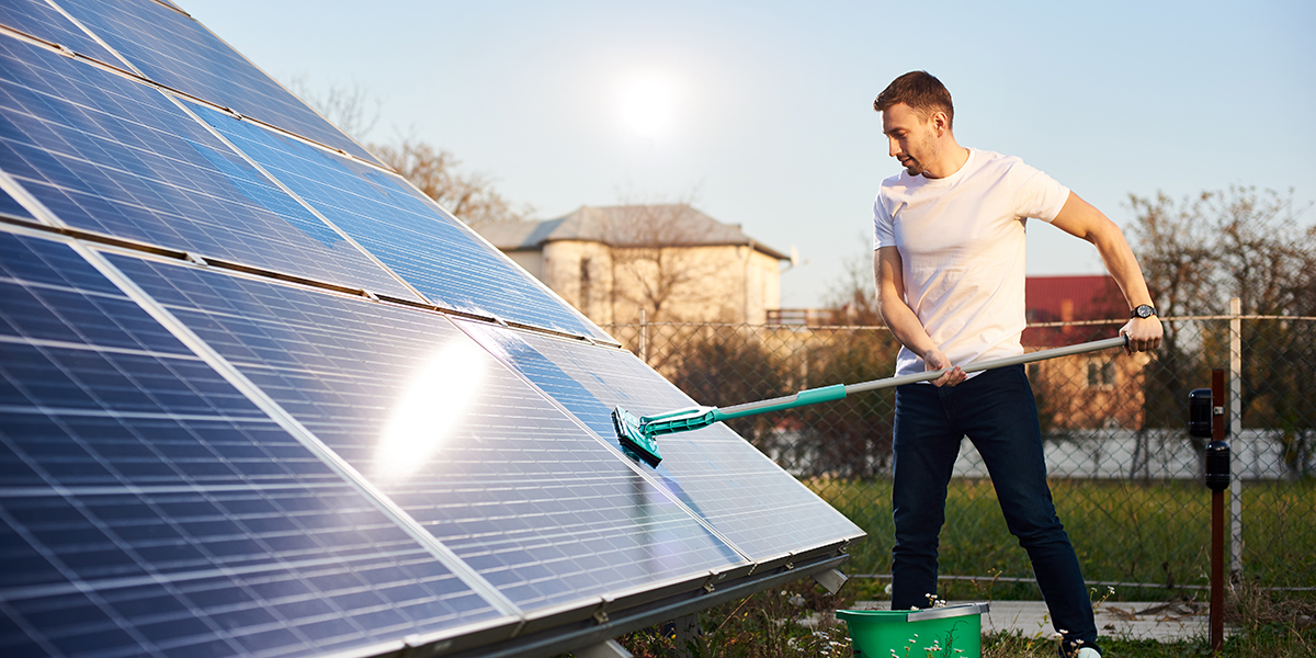 Young man is cleaning a solar panel with a mop, pv plant in rural area, side view